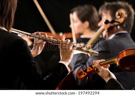 Professional female flutist in concert with symphony orchestra players on background.