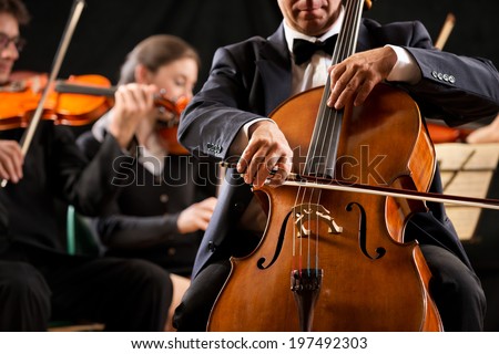 Cello professional player with symphony orchestra performing in concert on background.