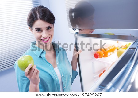 Young cheerful woman taking a green apple from refrigerator and smiling at camera.