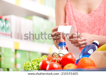 Unrecognizable woman checking a long supermarket receipt with grocery on foreground.