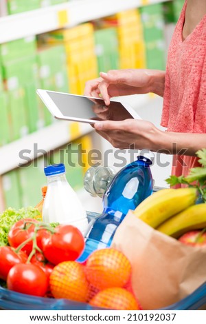 Young woman at supermarket shopping with digital tablet and full basket on foreground.