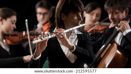 Female flutist close-up with orchestra performing on background.