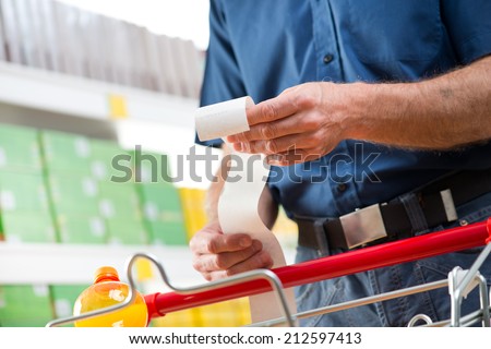 Man at supermarket examining a long grocery receipt with shopping cart on foreground.