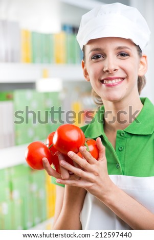 Young female sales clerk smiling and holding fresh tomatoes at supermarket.