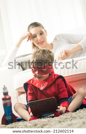 Cute superhero boy playing videogames sitting on carpet with his mother on background.