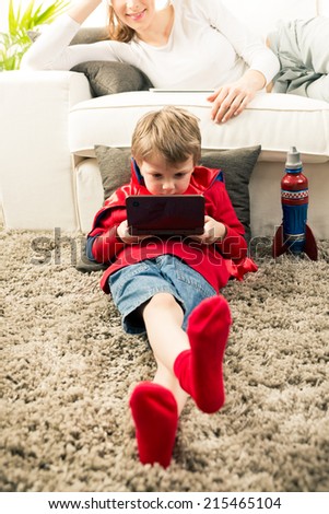 Little superhero boy playing videogames and mother lying down on sofa in the living room.