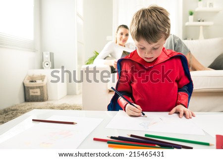 Cute superhero boy drawing with mother reading on background.