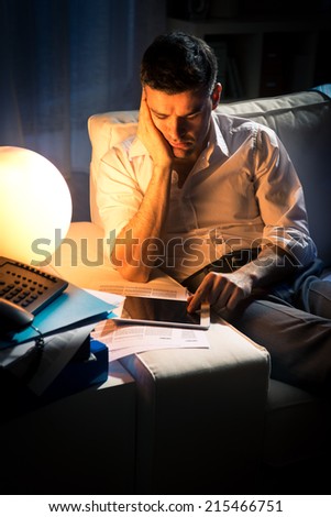 Exhausted businessman working late at night in the living room holding a tablet.