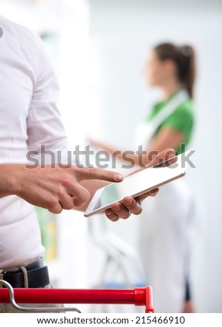 Customer using a digital tablet at supermarket with sales clerk on background.