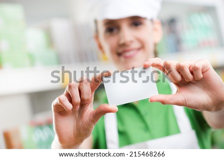 Female sales clerk holding a white card and smiling with supermarket shelf on background.