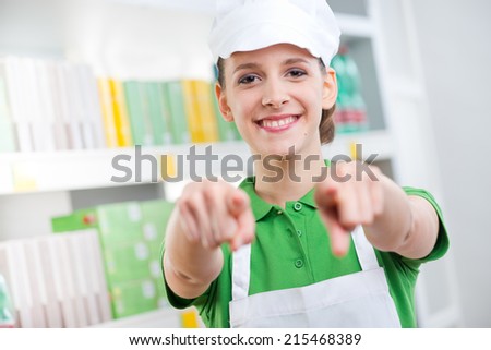 Young female sales clerk at supermarket smiling and pointing at camera.