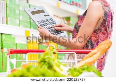 Woman shopping at supermarket with shopping cart and big calculator checking prices.