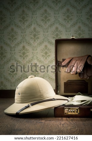 Explorer packing with open leather briefcase, pith hat, leather gloves on wooden surface.