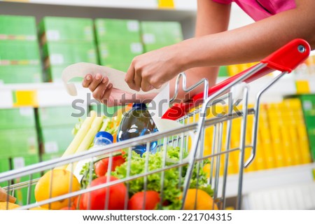 Unrecognizable woman checking a long grocery receipt leaning to a full shopping cart at store.