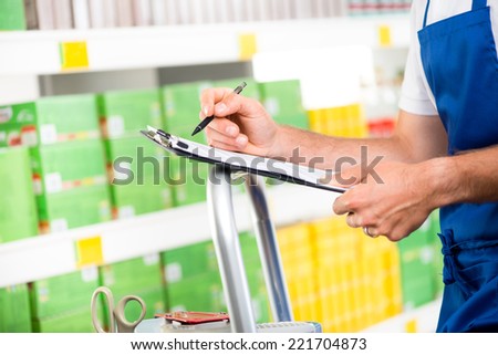 Supermarket clerk at work holding pen and clipboard with shelf on background, hands close-up.