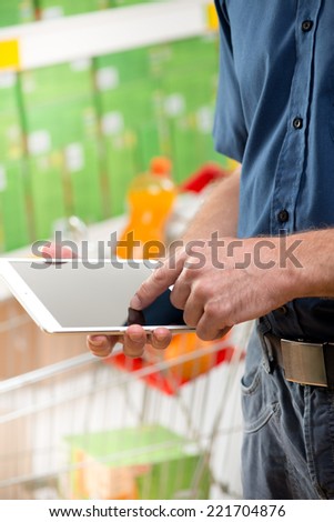 Man using digital tablet at supermarket with trolley and shelves on background.