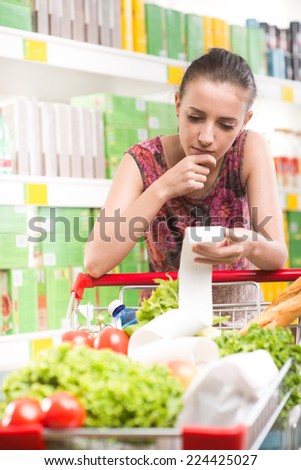 Young woman with hand on chin examining a grocery receipt at supermarket leaning to a shopping cart.
