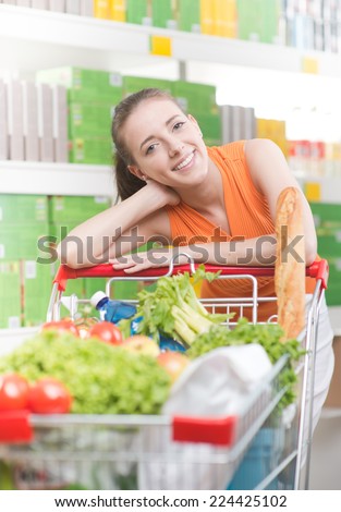 Smiling woman at supermarket with full shopping cart and shelves on background.