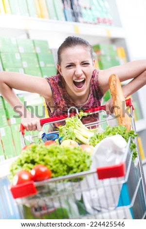 Angry woman pushing a full shopping cart at store with shelves on background.