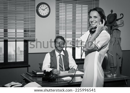 Young 1950s secretary answering phone calls in director's office and smiling.