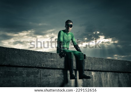 Green pensive superhero sitting on a concrete wall with dramatic cloudy sky on background.