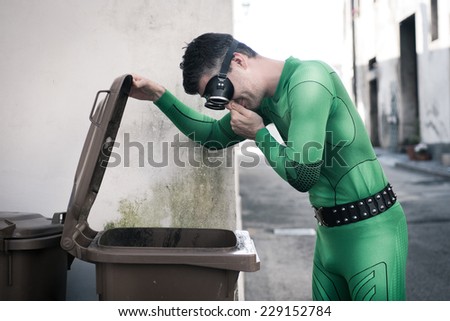 Green superhero holding his nose and opening a trash bin in the street.