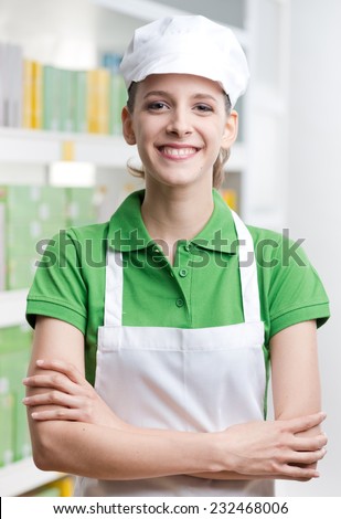 Young female sales clerk with crossed arms smiling at camera with supermarket shelf on background.