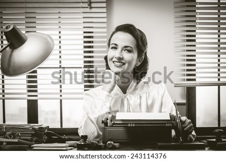 1950s style secretary working at office desk and smiling with hand on chin