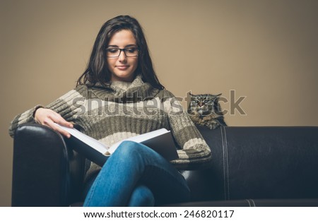 Young relaxed woman on sofa reading a book with curious cat peeking from back