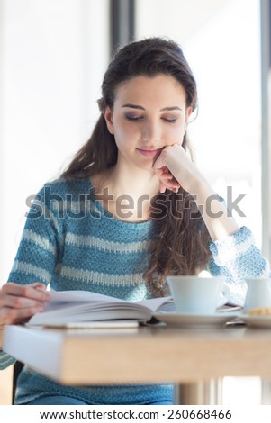 Cute young girl student at the cafe reading a book and sitting at the table