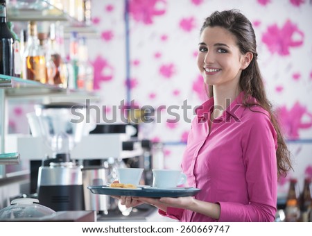 Young smiling waitress in pink shirt serving coffee at the bar, floral wallpaper on background