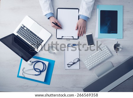 Professional doctor writing medical records on a clipboard with computer and medical equipment all around, desktop top view