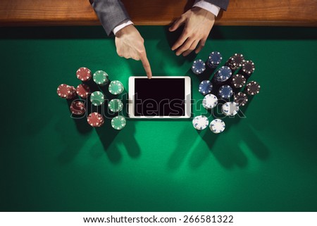 Male elegant player touching a digital tablet screen with chips all around on green table, top view