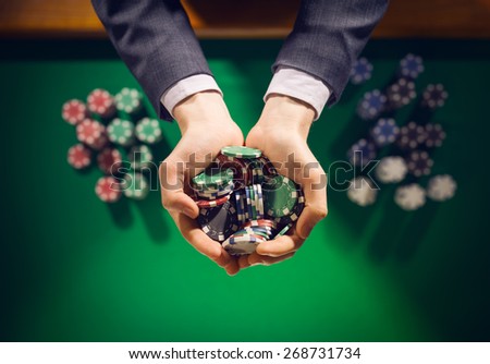 Elegant male casino player holding a handful of chips with green table on background, hands close up top view