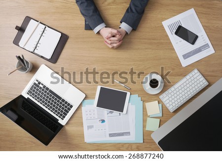 Confident businessman sitting at his office desk with hands clasped, paperwork and computers around, hands detail top view