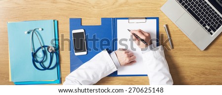 Doctor working at office desk writing medical records on a clipboard hands close up top view