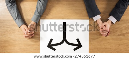 Business man and business woman sitting at desk with hands clasped and a sign with two arrows at center, separation and divorce concept, hands top view, unrecognizable people