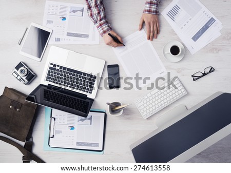 Businessman working at office desk and signing a document, computers and paperwork all around, top view