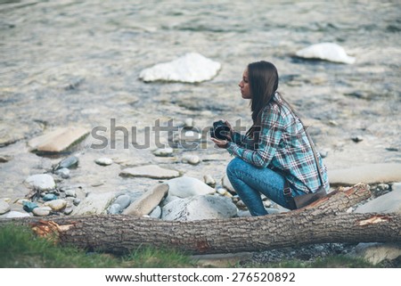 Young tourist woman exploring nature and holding a digital camera, she is sitting along the river