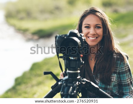 Smiling female photographer using a professional digital camera on a tripod, natural landscape on background