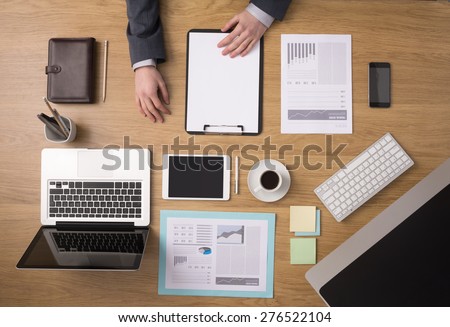 Businessman working at office desk holding a clipboard with a blank sheet, paperwork and computers around, top view