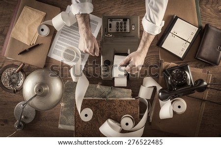 Messy vintage accountant\'s desktop with adding machine and paper rolls, he is working with the calculator