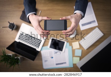 Businessman working at office desk hands top view with laptop and financial reports: he is using a mobile touch screen smart phone