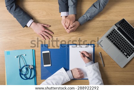 Doctor writing down medical records and talking with a couple during a visit, desktop top view, unrecognizable people