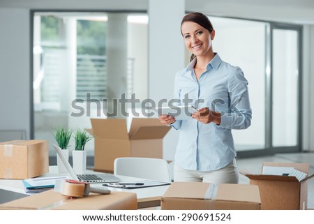 Confident smiling business woman moving in her new office, she is unpacking boxes and using a digital tablet