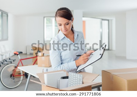 Smiling businesswoman writing a relocation checklist for her office on a clipboard and looking into an open carton box