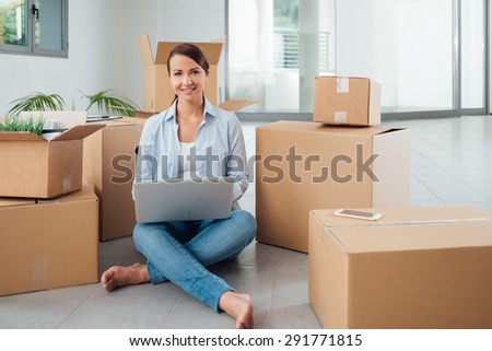Beautiful woman moving in her new house and unpacking, she is sitting on the floor surrounded by boxes, using a laptop and smiling at camera