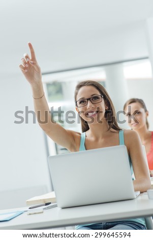 Smart smiling girl sitting at school desk and raising her hand to ask or answer a question