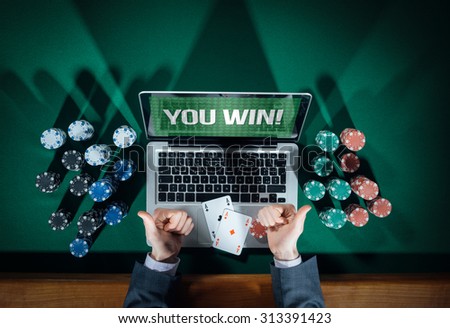 Man thumbs up playing online poker with laptop on a green table with chips all around, top view