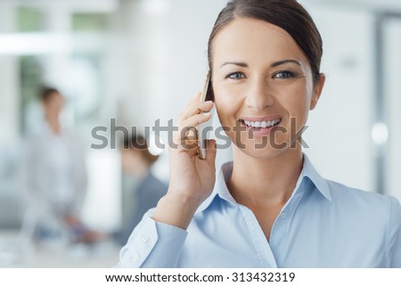 Smiling confident business woman holding a smart phone and calling, office interior and business people on background, selective focus